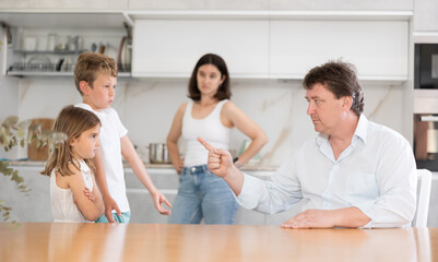 Fototapeta premium At home in kitchen, mom and dad scold young children for imprudent act. Husband and wife scold children, son denies his bad behavior. Children with guilty look stand in front of angry father