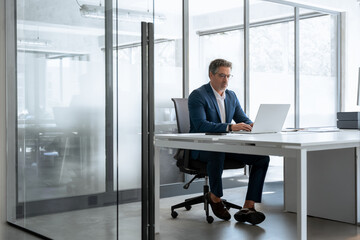 Mature 50s Latin business man ceo manager using laptop computer in office. Focused senior businessman entrepreneur sitting at desk working typing looking at screen. Full body portrait. Copy space