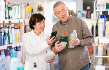 Couple elderly man and woman buyers scanning qr code for dishwashing detergent in household chemicals store