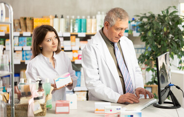 Fototapeta premium Elderly experienced pharmacist works at computer in a drugstore. Young assistant helps to find pills
