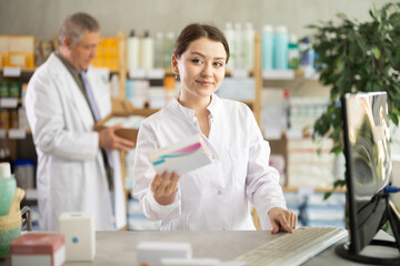 Obraz premium Girl pharmacist uses computer at workplace. Employee reads regulations and laws, makes report, reads information about drugs. Male colleague work in background