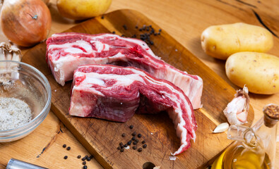 Raw beef ribs lie next to vegetables and spices - process of cooking homemade festive dish. Unpeeled vegetables, spices, olive oil and cooking utensils are placed on table before processing ingredient