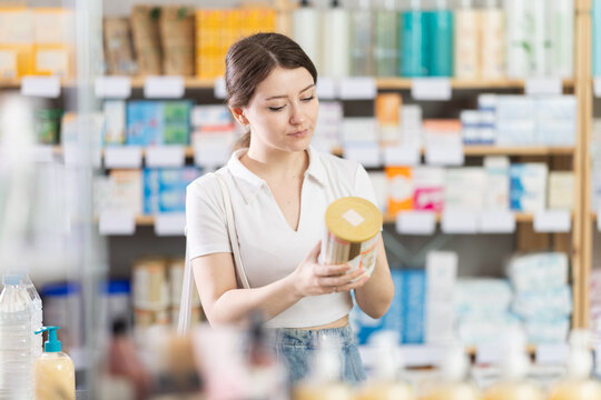 Thoughtful young woman choosing baby formula in pharmacy, reading label on container and examining ingredients before purchase