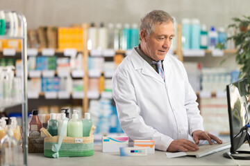 Confident professional senior pharmacist in white coat working behind drugstore counter, inputting...