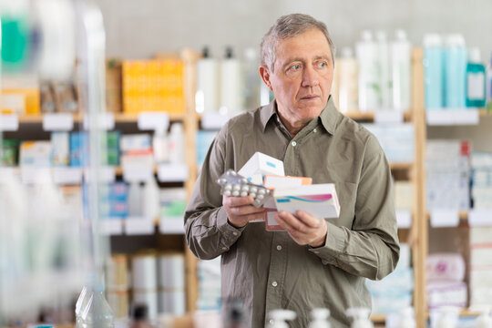 Senior man in pharmacy looking overwhelmed while holding several boxes of pills and blister packs, trying to choose right medicine