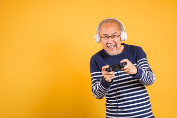 Portrait Asian smiling old man with headphones holding game controller studio shot isolated yellow background, senior man pensioner feeling winning playing a video game with joystick console © sorapop