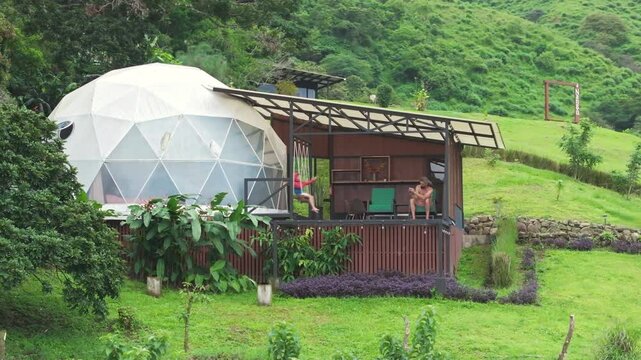 A father and son relax on the veranda of a geodesic dome, enjoying the breathtaking natural beauty of Costa Rica, with an aerial view highlighting the serene atmosphere around them
