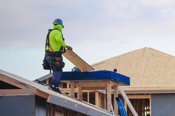 A construction worker in high-visibility gear installs roof sheathing on a sloped structure as part...