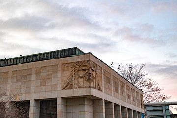Fototapeta premium Stone Relief of a Solemn Face on a Modern Building Facade at Dusk with Birds on Rooftop and Bare Tree Silhouette