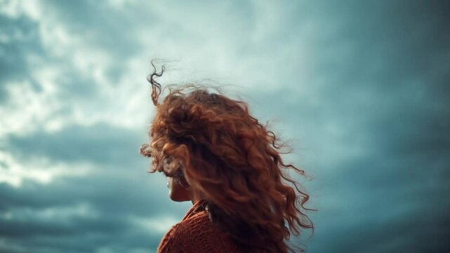 Curly-haired woman stands as wind lifts hair under dramatic clouds. Slow arc shot. 4K.
