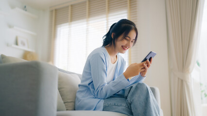 Obraz premium Smiling pretty young Asian woman sitting on cozy couch, using funny mobile apps in living room. Woman at Home, Doing Online Shopping, Messaging Friends, Posting on Social Media
