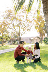Smiling parents squatting and looking at little girl in sunny park