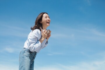 A cheerful Asian woman enjoying a sunny day at the beach. She is wearing a casual shirt and jeans standing in front of the blue sky background