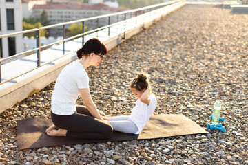 Mother and preteen daughter doing sit-ups on rooftop during sunny day. Woman helps child develop strength and form through guided exercise session.