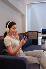 Fototapeta premium Young woman relaxing on sofa while listening to music with headphones and browsing songs on smartphone.