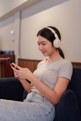 Young woman relaxing on sofa while listening to music with headphones and browsing songs on smartphone.