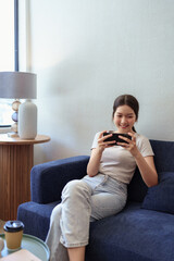 Young woman enjoying mobile gaming on smartphone while sitting on sofa in cozy daylight cafe corner