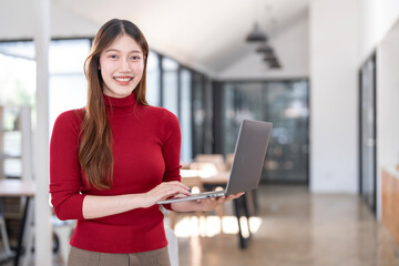 Woman in red sweater holding laptop computer typing on keyboard standing at office, looking at camera,