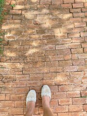 A top-down view of a woman's feet, clad in light-colored, white or cream, slip-on shoes, standing on an outdoor path. There are som faint, dappled shadows on the path, filtered sunlight. A rustic walk