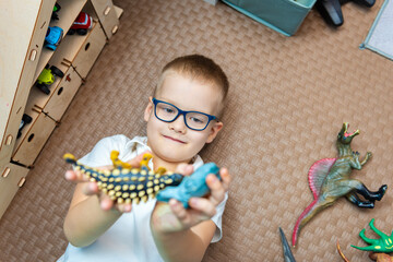 A boy with glasses plays with dinosaurs lying on his back in the playroom. View from above. Selective attention to your favorite toys, the choice of toys.