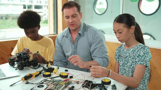 Smart mentor teaching about electronic equipment while fixing car model. Caucasian teacher pointing while explain robotic construction at table with laptop, car model scatter around. Edification