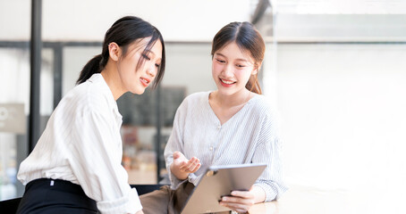 Two asian business women sit at desk consulting discuss project details together at office, using digital tablet