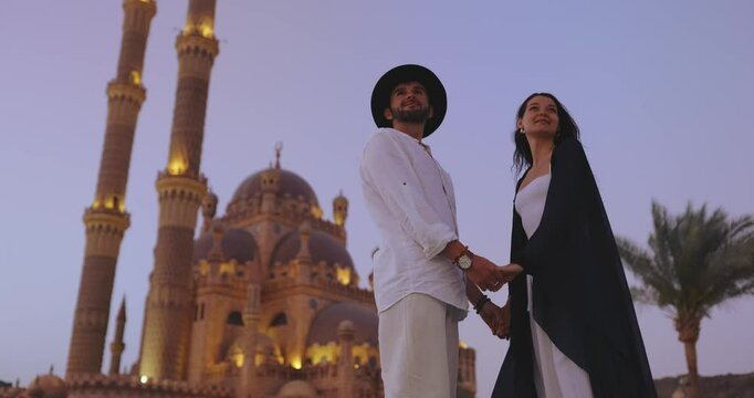 A joyful couple raises their hands in celebration in front of the beautifully illuminated Sahaba Mosque at twilight in Sharm El Sheikh, Egypt. Ideal for high-quality travel, cultural, and romantic