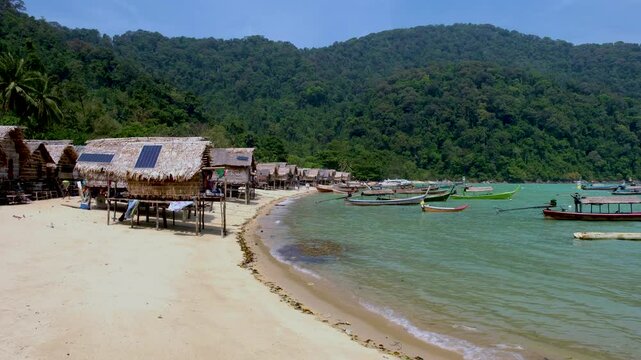 Wander through a tranquil coastal village in Koh Surin Thailand featuring stilted huts along a sandy beach. Vibrant fishing boats bob on the crystal-clear water, surrounded by lush greenery.