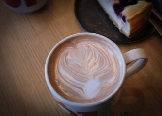 Cup of Cappuccino coffee with latte art, served on a wooden table in a restaurant with cheesecake on the side