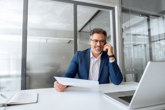 Positive financial specialist senior man working on laptop computer in office, having cell phone call. Mature businessman professional using pc, analysing business project, holding report document