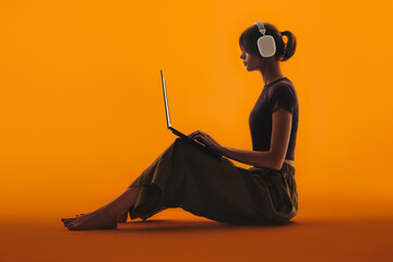 Young woman with headphones sitting on the floor in the studio and working on a laptop. Orange background.