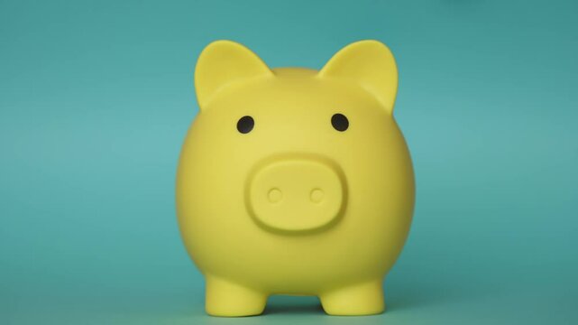 Hand inserting golden coin into bright yellow piggy bank, representing strategic financial planning and personal wealth accumulation against vibrant turquoise background