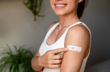 Smiling woman pointing at adhesive bandage on her upper arm at vaccination site after injection