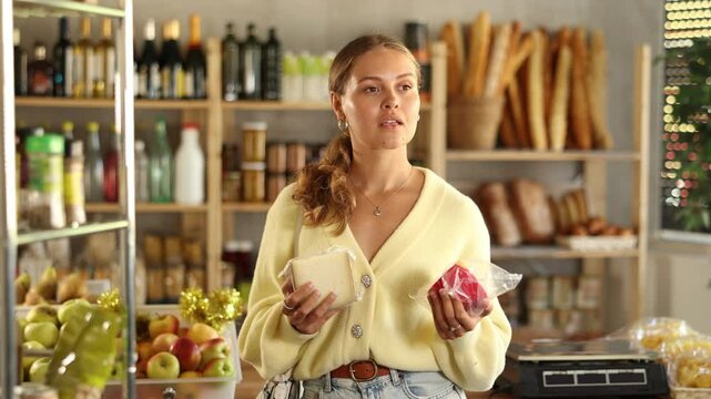 Young woman with cheese in supermarket with food shelves in the background. Interested buyer chooses hard cheeses in a grocery store
