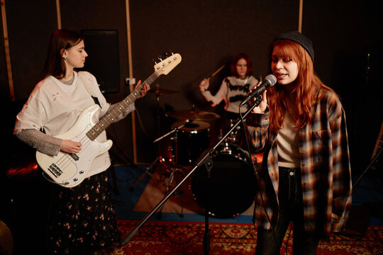 Medium full shot of young redhead girl singing into microphone playing new song with girl rock band in recording studio with dim light