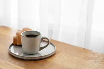 Cup of freshly brewed coffee and croissant on a plate on a wooden table