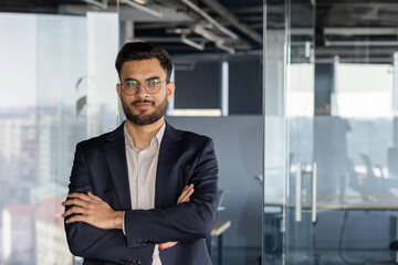 A smiling businessman with arms crossed, standing in an office with a window background. He is...