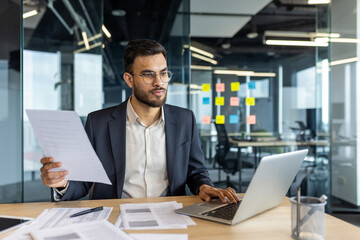 A focused businessman in glasses reviews documents while working on a laptop at his desk. The office setting features glass walls and sticky notes.