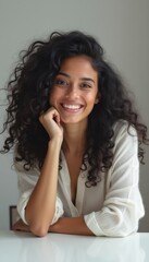 A joyful young woman with voluminous curly hair leans on a white surface, exuding confidence and warmth. Her bright smile and relaxed pose create an inviting atmosphere, perfect for showcasing beauty