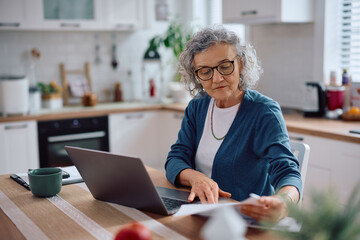 Senior woman analyzing home finances while using laptop in kitchen.