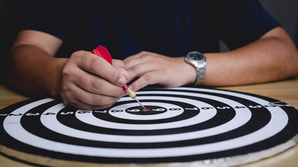 A hand holding a red dart aimed precisely at the bullseye of a black-and-white dartboard, symbolizing focus, accuracy, and achievement.