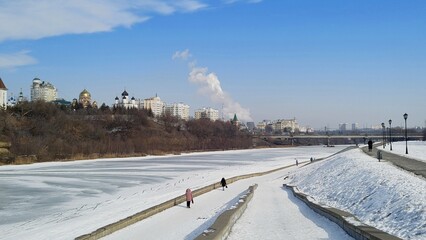 Orel city embankment in winter. Frozen Oka River. People walking along shore. White snow and bright blue sky. Urban landscape. Beautiful Russian city