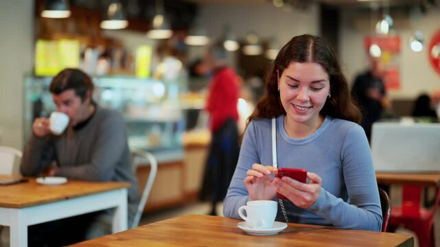 Young woman in cafe drinks coffee and using mobile phone, scrolls through social media feed, chats online, calls friend, talks on phone. Free Wi-Fi zone, Internet cafe.