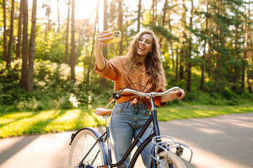 Selfie time! Young woman riding a bicycle with a phone taking a selfie in the park. Happy woman...