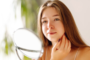 Beautiful woman with shining clear skin looking at the mirror in a cozy room. Young woman enjoying...