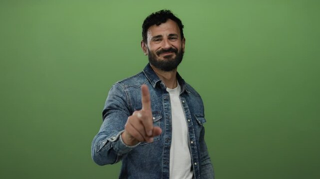 Hispanic man in denim shirt with beard making no gesture with finger against green background.