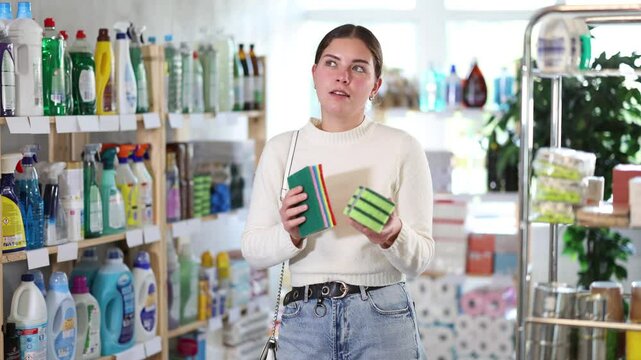 Young woman choosing cleaning sponges for dishes and surfaces. Customer in the household section of a supermarket buys sponges of different hardness