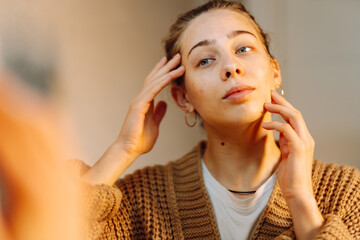 Portrait of a young woman touching her face and looking at the mirror in a cozy room. Beautiful...