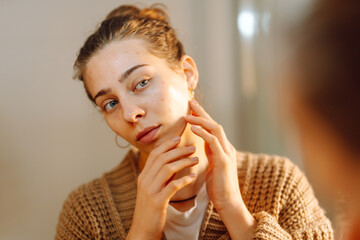 Portrait of a young woman touching her face and looking at the mirror in a cozy room. Beautiful...