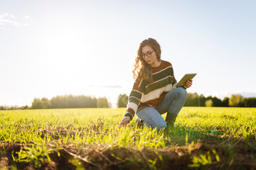 Young female farmer with a digital tablet inspects young plants in an agricultural field. Female...